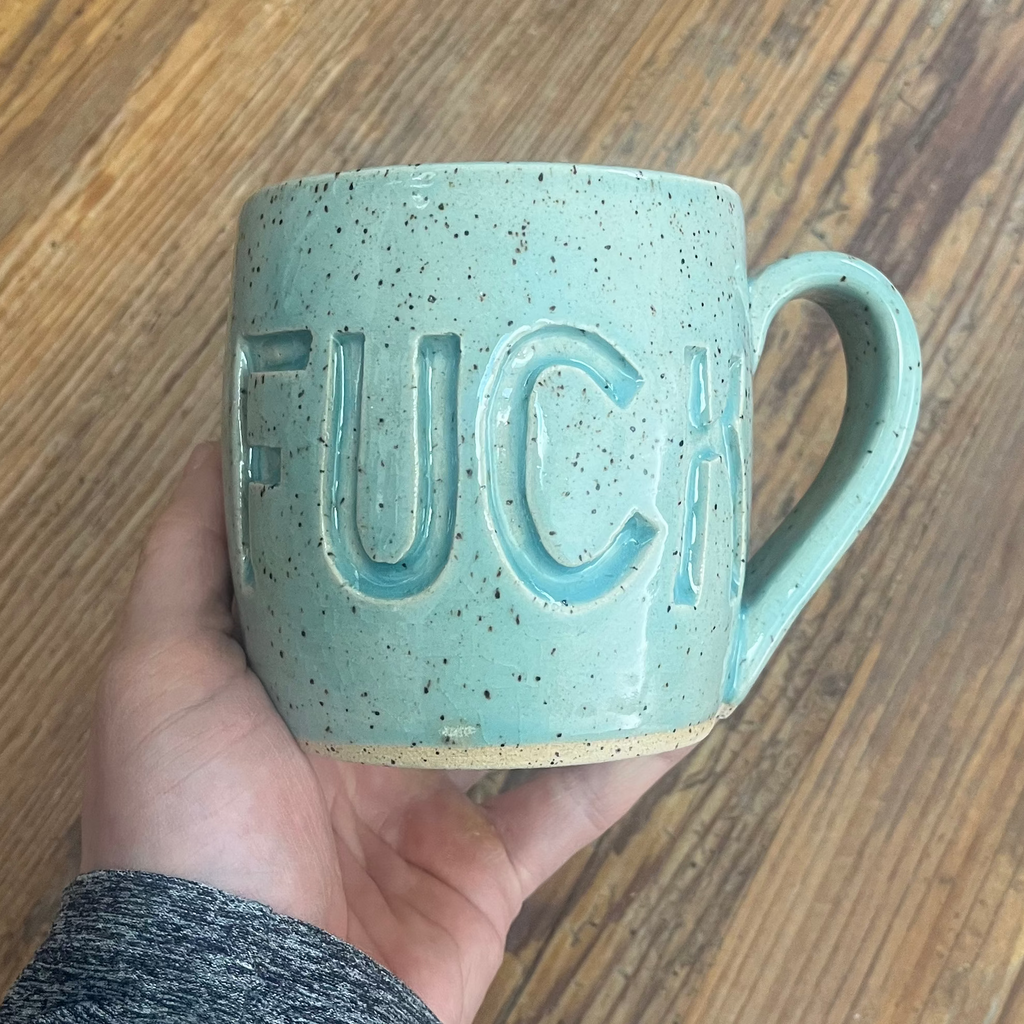 Hand holding an aqua mug with fuck carved into it.