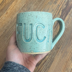 Hand holding an aqua mug with fuck carved into it.
