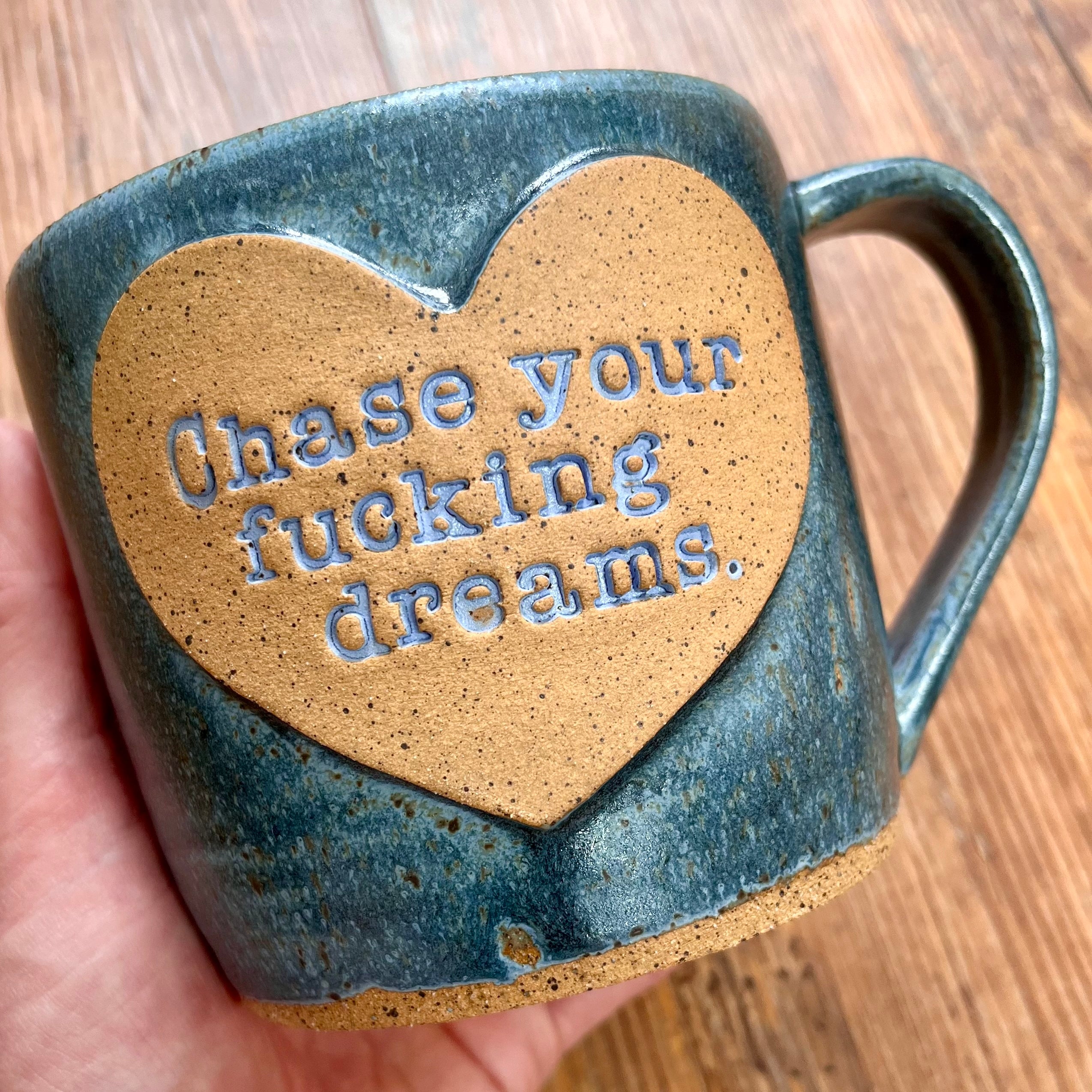 A greenish blue mug with the phrase "chase your fucking dreams" inside a brown clay heart