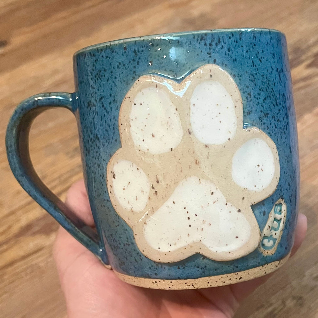 A ceramic mug with a speckled blue glaze and a large raised cream-colored paw print on the front with the name "Gus" on it, held in a hand over a wooden surface.