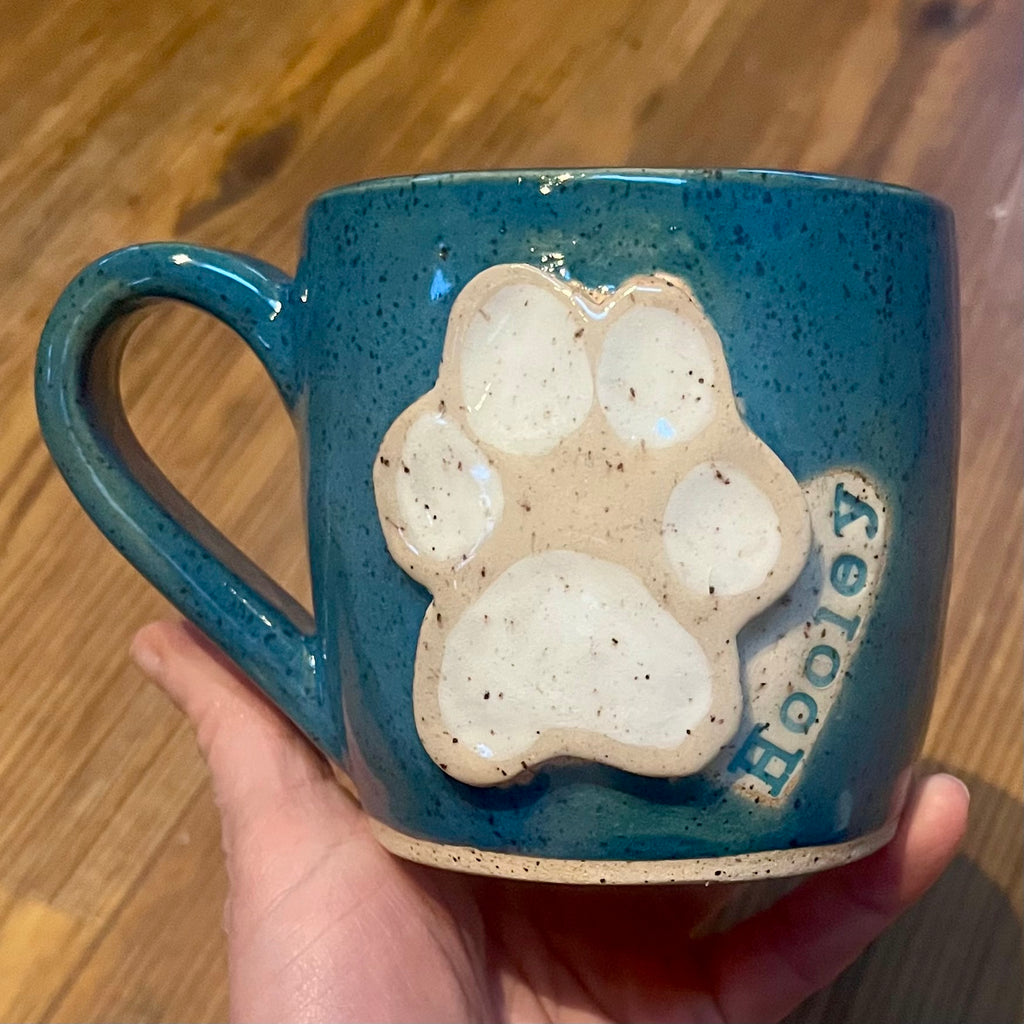 A hand holds a speckled teal ceramic mug with a raised cream-colored paw print and the name 'Hooley' on the side, set against a wooden background.