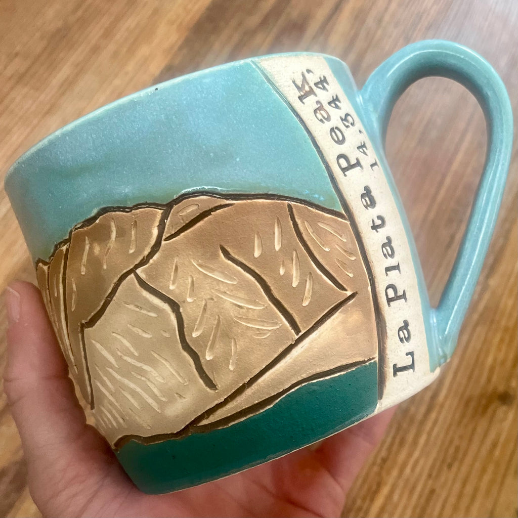 A handmade ceramic mug held in a hand over a wooden table. The mug features a turquoise blue and dark teal glaze with a raised relief design of the Colorado 14'er La Plata Peak, and includes the name and elevation of the peak 