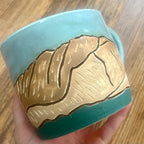 A handmade ceramic mug held in a hand over a wooden table. The mug features a turquoise blue and dark teal glaze with a raised relief design of the Colorado 14'er La Plata Peak 