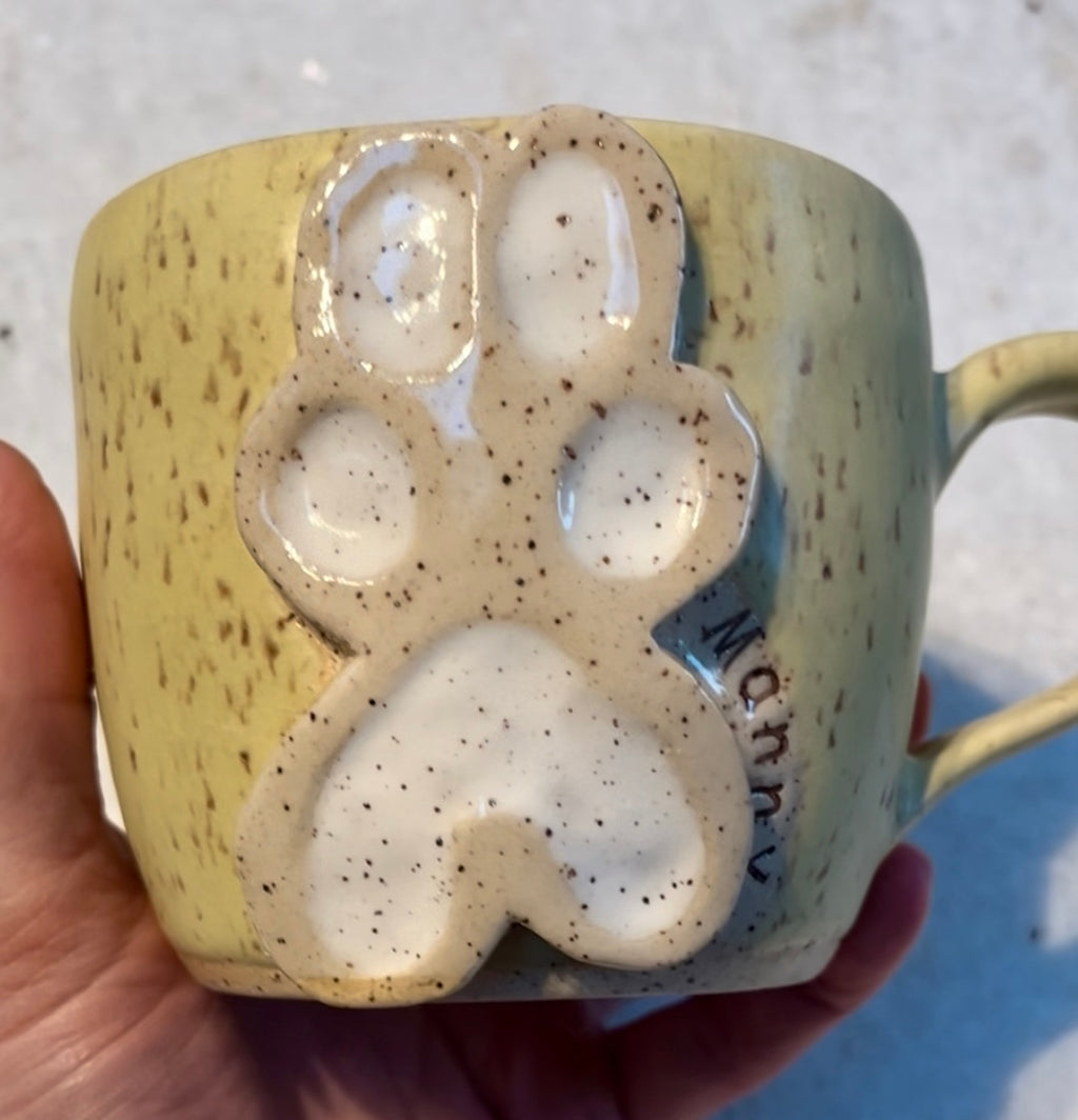 A handmade ceramic mug held by a hand, featuring a textured speckled yellow-green glaze with a raised white and brown paw print design on the front and the name 'Manny' embossed on the side.