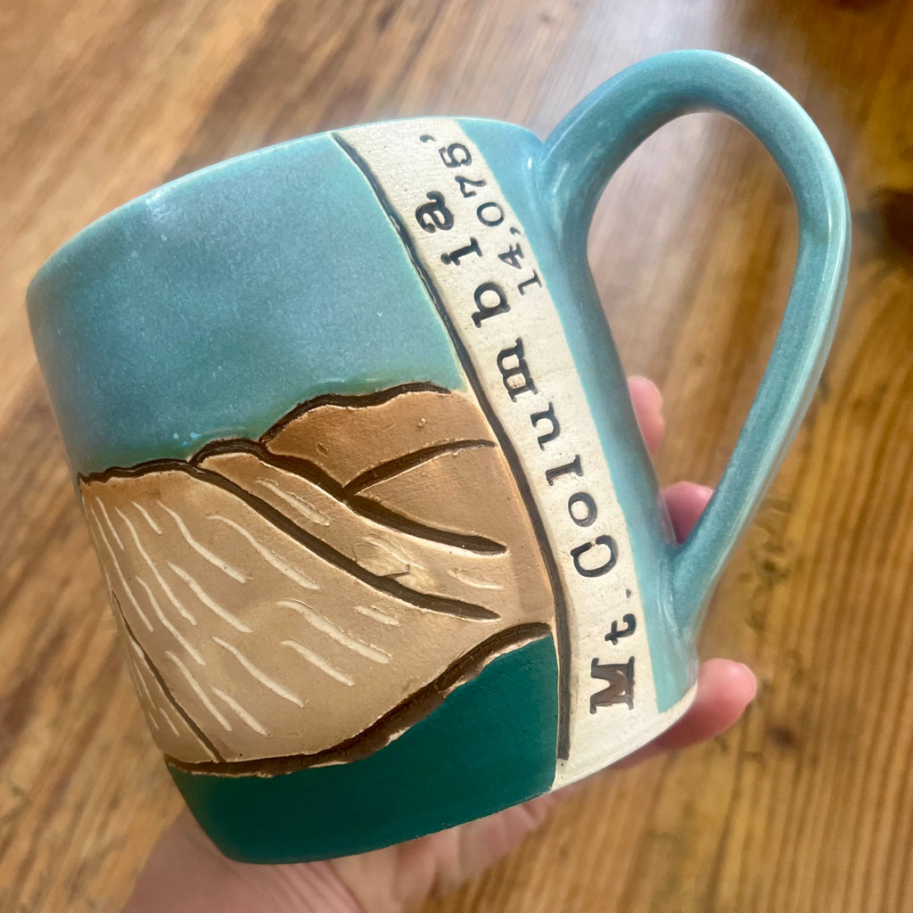 A handmade ceramic mug held in a hand over a wooden table. The mug features a turquoise blue and dark teal glaze with a raised relief design of the Colorado 14'er Mount Columbia and a vertical banner inscribed with 'Mt. Columbia 14,075'