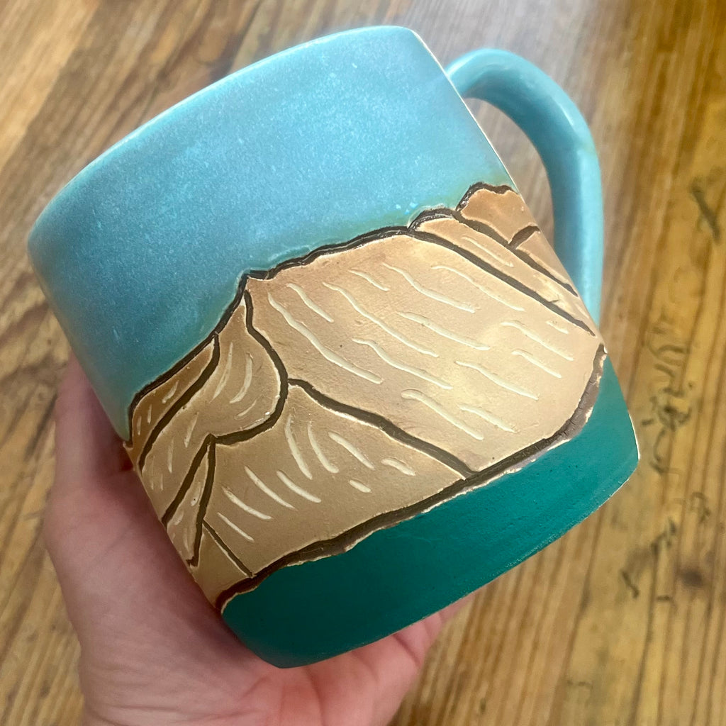 A handmade ceramic mug held in a hand over a wooden table. The mug features a turquoise blue and dark teal glaze with a raised relief design of the Colorado 14'er Mount Columbia 