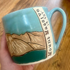 A handmade ceramic mug held in a hand over a wooden table. The mug features a turquoise blue and dark teal glaze with a raised relief design of the Colorado 14'er Mount Massive 