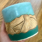 A handmade ceramic mug held in a hand over a wooden table. The mug features a turquoise blue and dark teal glaze with a raised relief design of the Colorado 14'er Mount Massive 