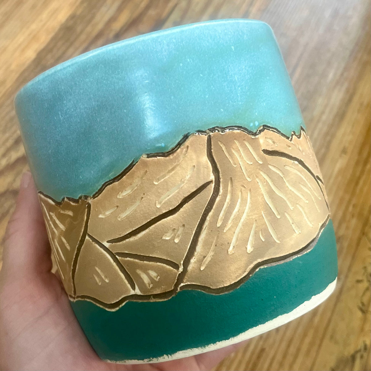 A handmade ceramic mug held in a hand over a wooden table. The mug features a turquoise blue and dark teal glaze with a raised relief design of the Colorado 14'er Mount Massive 