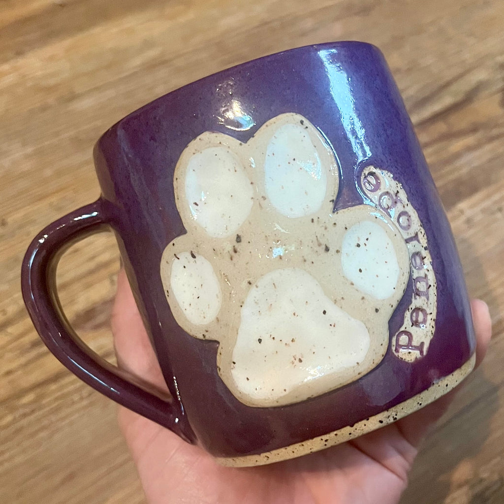 A hand holds a purple ceramic mug with a large beige paw print and the Penelope on the side, set against a wooden background.
