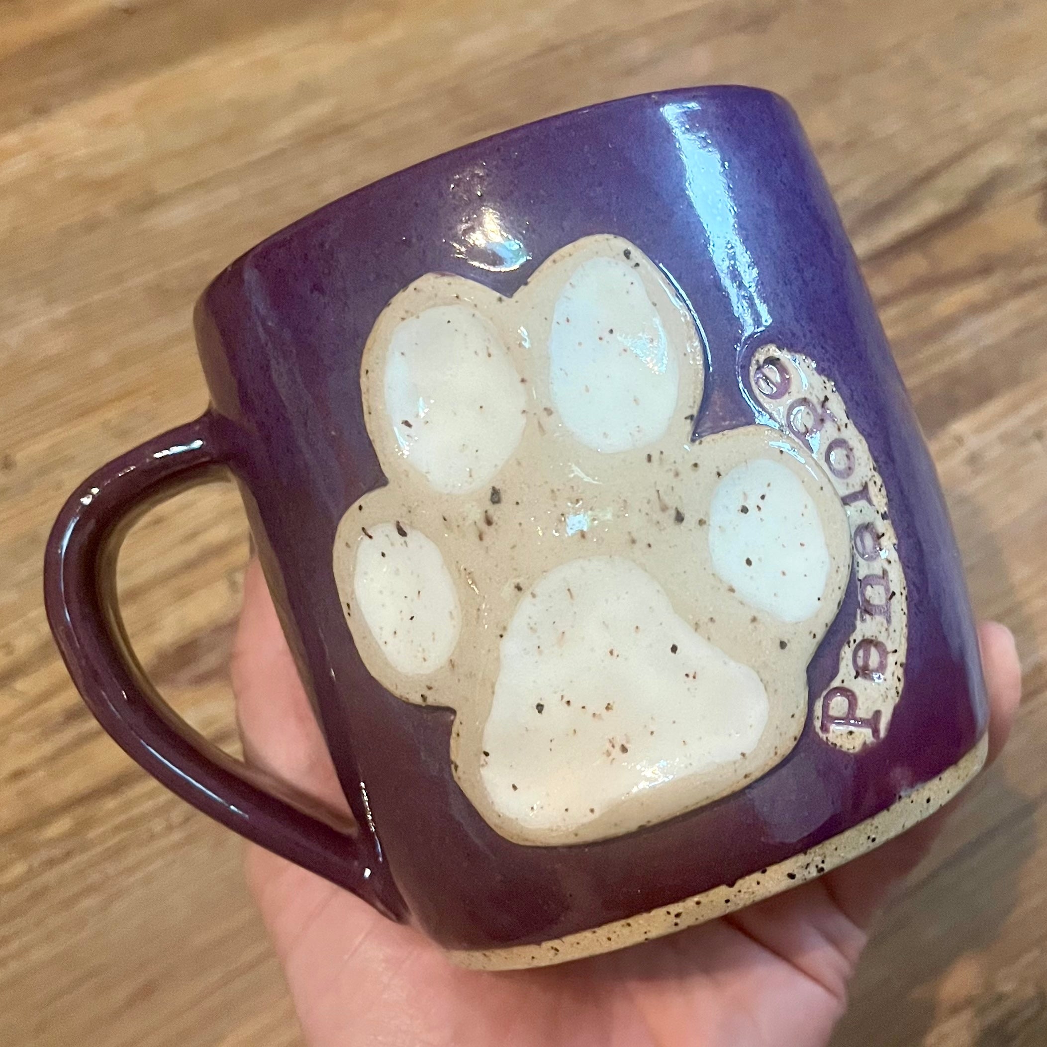 A hand holds a purple ceramic mug with a large beige paw print and the Penelope on the side, set against a wooden background.