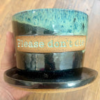 A handmade ceramic planter with an attached saucer, held by a hand against a wooden background. The planter features a glossy black and teal glaze with a textured tan band in the middle containing the raised text 'Please don't die'.