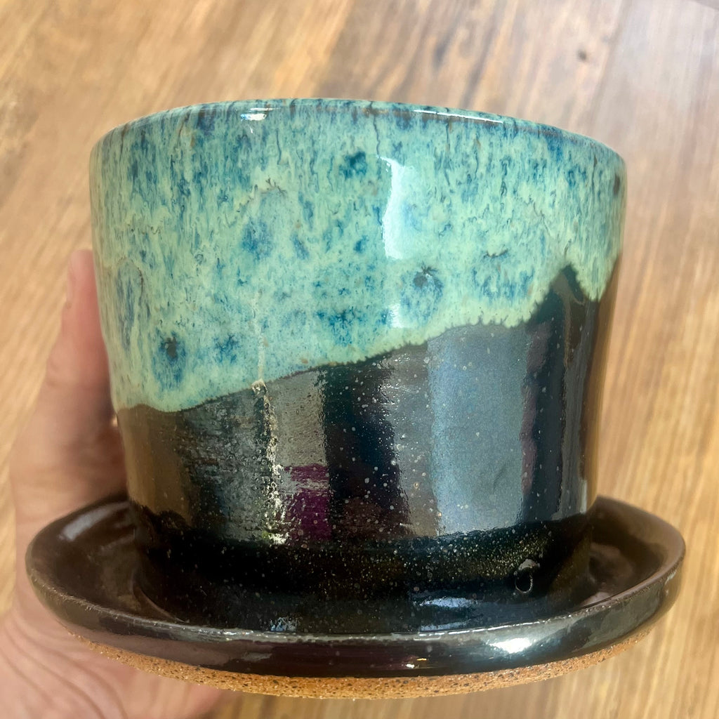 A handmade ceramic planter pot with an attached saucer, held by a hand against a wooden background. The pot features a glossy glaze with a textured light blue-green top half and a dark black-brown bottom half.