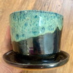A handmade ceramic planter pot with an attached saucer, held by a hand against a wooden background. The pot features a glossy glaze with a textured light blue-green top half and a dark black-brown bottom half.