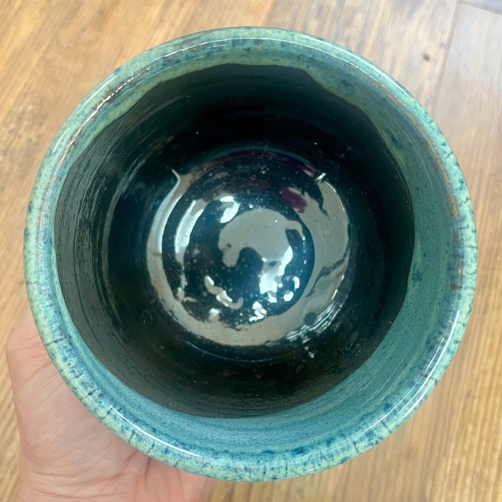 A handmade ceramic planter with a glossy black interior and a textured light teal glazed rim, held by a hand over a wooden table background.
