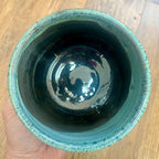 A handmade ceramic planter with a glossy black interior and a textured light teal glazed rim, held by a hand over a wooden table background.