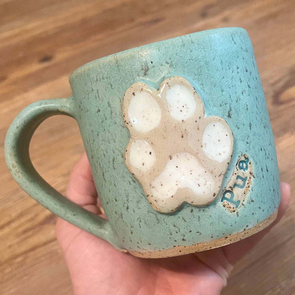 A handmade ceramic mug with a speckled sage green glaze, featuring a raised beige paw print design on the side and the name 'Pua' in blue lettering, held in a person's hand over a wooden table background.