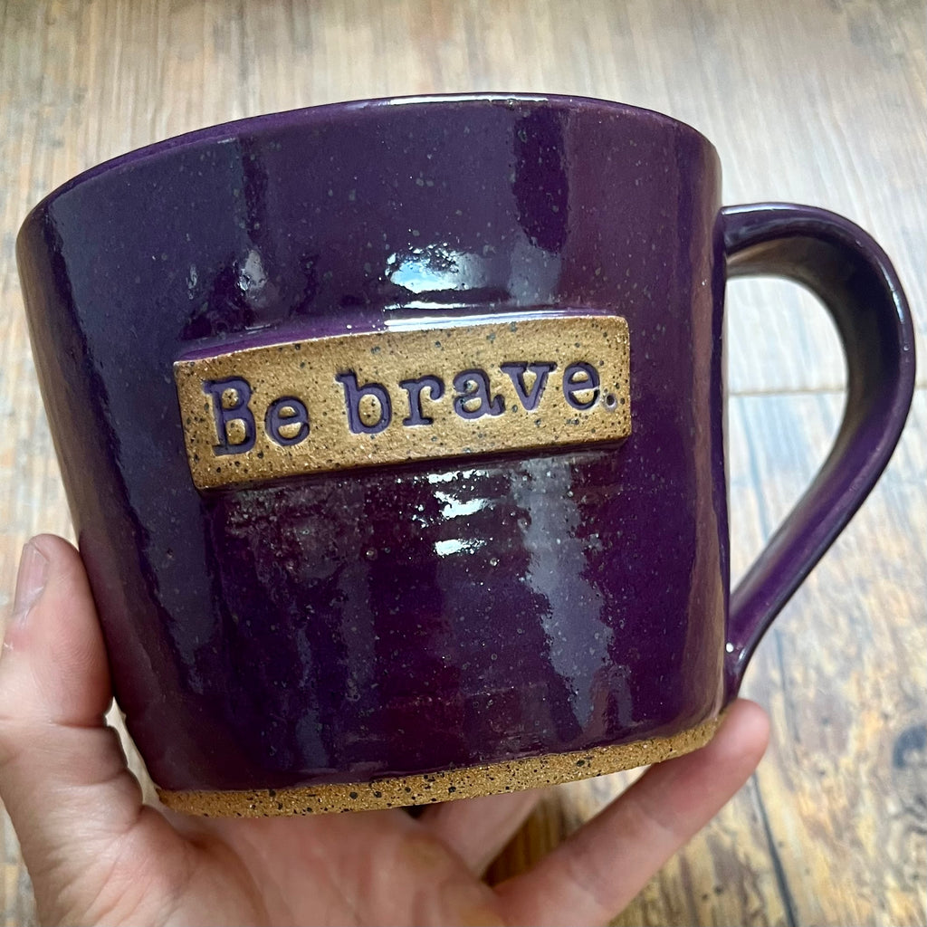 a purple mug featuring the phrase "be brave" in a clay rectangle