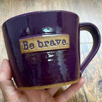 a purple mug featuring the phrase "be brave" in a clay rectangle