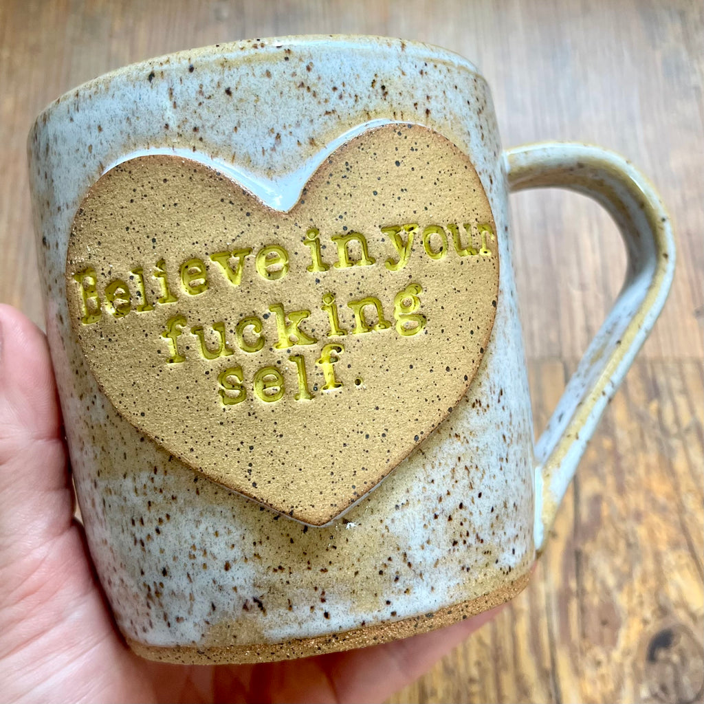 a white mug featuring the phrase "Believe in your fucking self" in a clay heart