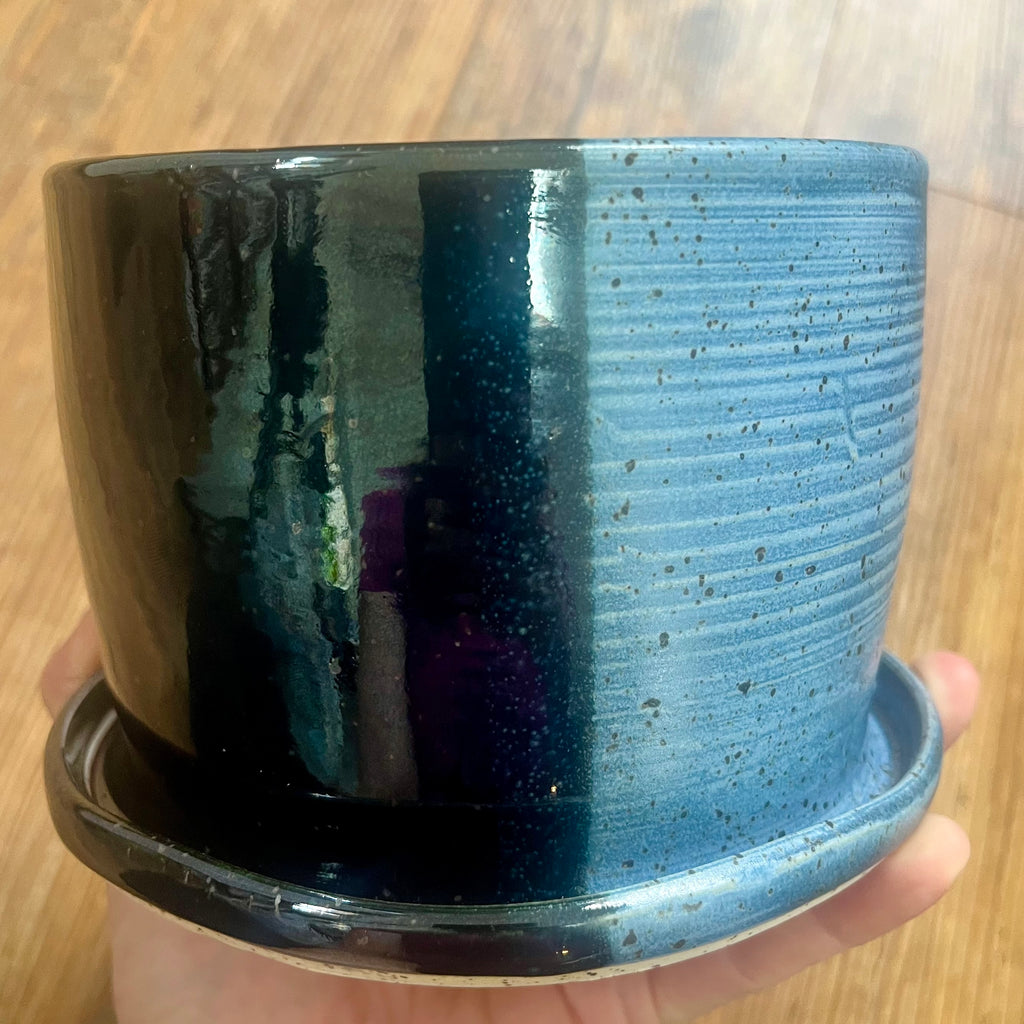A handmade ceramic planter pot with an attached saucer, held in a hand against a wooden floor background. The pot features a glossy black left side and a textured, ribbed blue-gray right side with dark speckles.