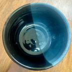 A handmade ceramic planter with a glossy glaze, shown from a top-down perspective on a wooden surface. The glaze features a distinct color split: a deep, dark black on one side and a rich, dark teal-blue on the other.