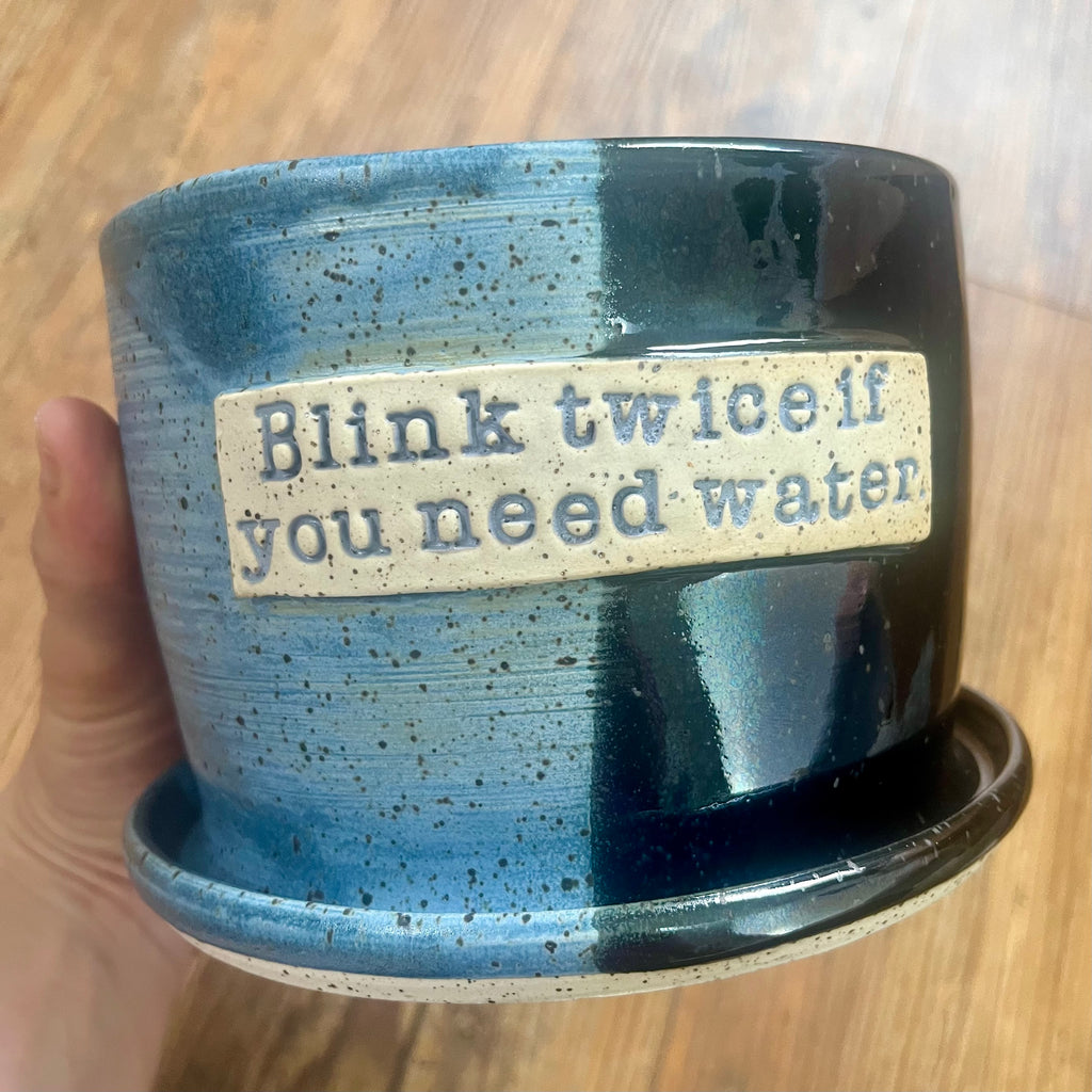 A handmade ceramic planter pot with an attached saucer, held by a hand. The pot features a split glaze design of blue and black with dark speckles, and a beige label on the front with the text 'Blink twice if you need water'.