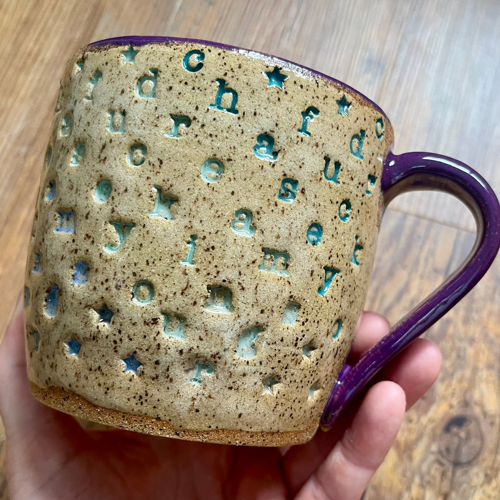 a mug with "chase your fucking dreams" stamped in diagonal stripes with a purple handle