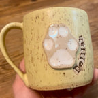 A handmade ceramic mug with a speckled light yellow glaze, featuring a raised beige paw print and the name 'Delilah' embossed on the side, held in a hand over a wooden surface.
