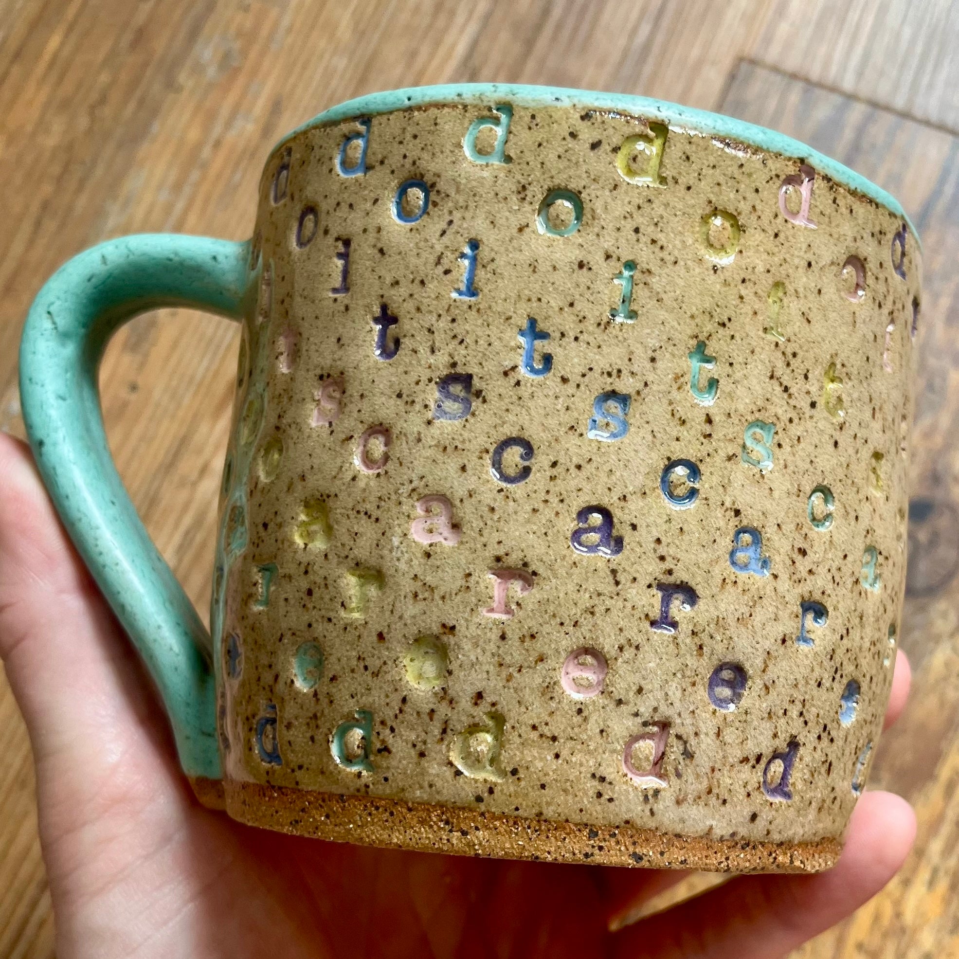 A mug with "do it scared" stamped in diagonal rainbow-colored stripes with a seaglass colored handle