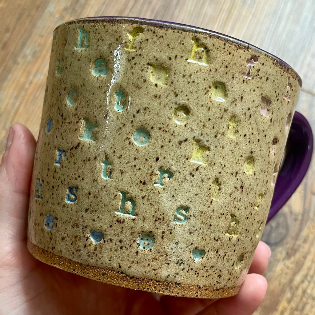 A handmade ceramic mug with a speckled beige and brown glaze, featuring raised letter shapes in light blue and yellow-green, held by a hand over a wooden background.