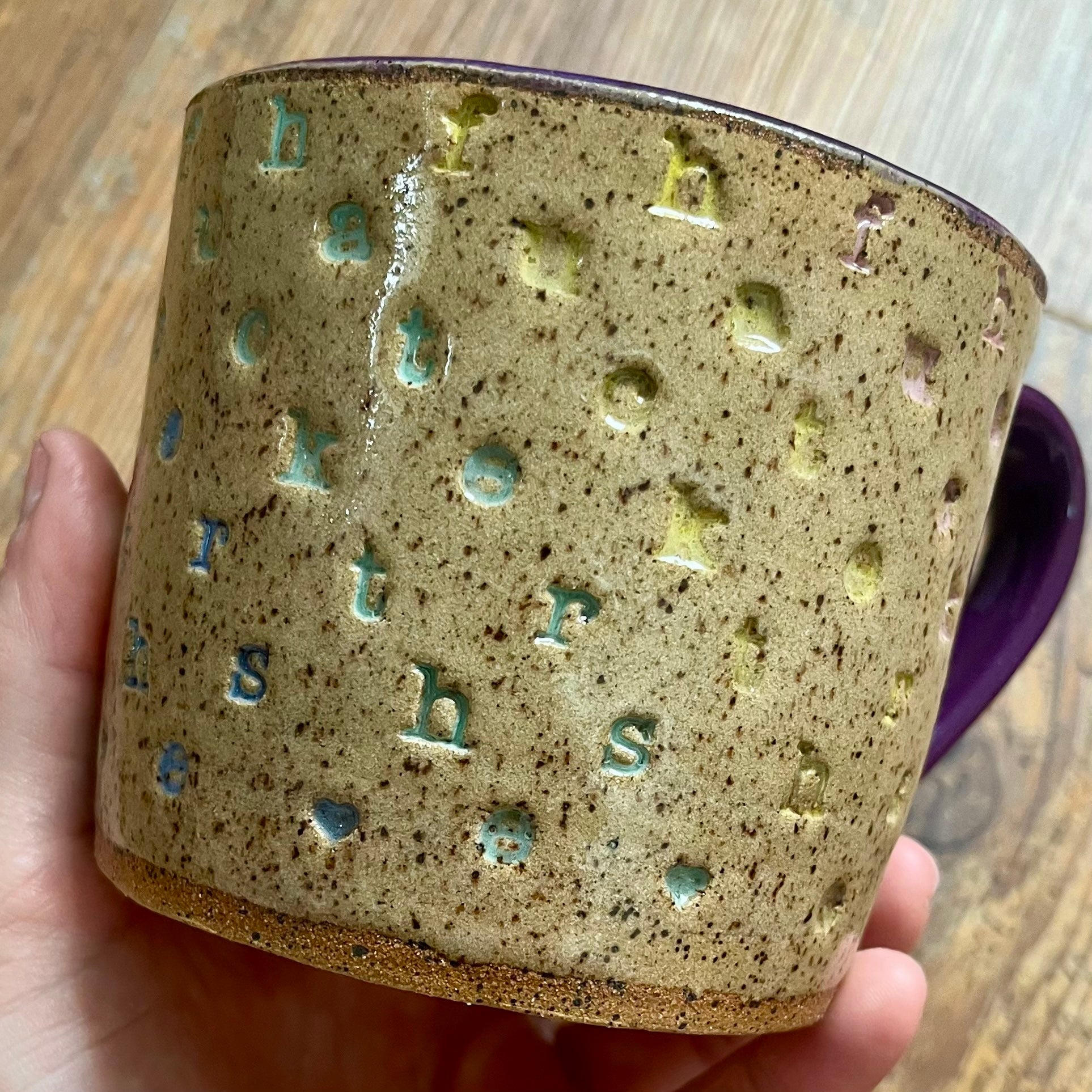 A handmade ceramic mug with a speckled beige and brown glaze, featuring raised letter shapes in light blue and yellow-green, held by a hand over a wooden background.