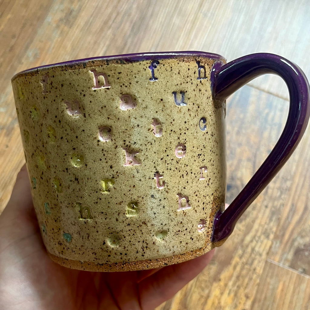 A handmade ceramic mug with a speckled beige and brown glaze, featuring embossed lowercase letters in various colors, held in a hand over a wooden table background.