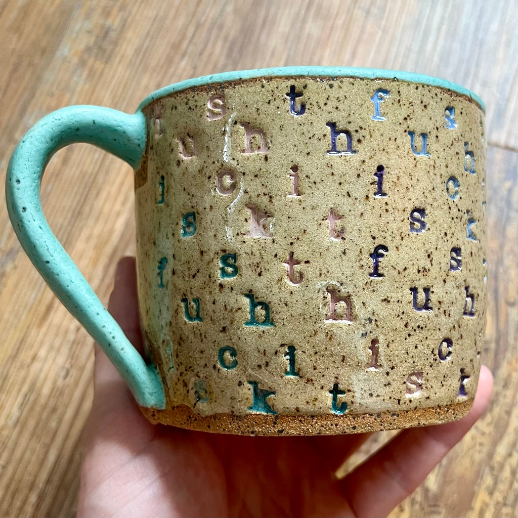 A mug stamped with "f*ck this sh*t" in diagonal stripes with a seaglass colored handle