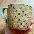 A mug stamped with "f*ck this sh*t" in diagonal stripes with a seaglass colored handle