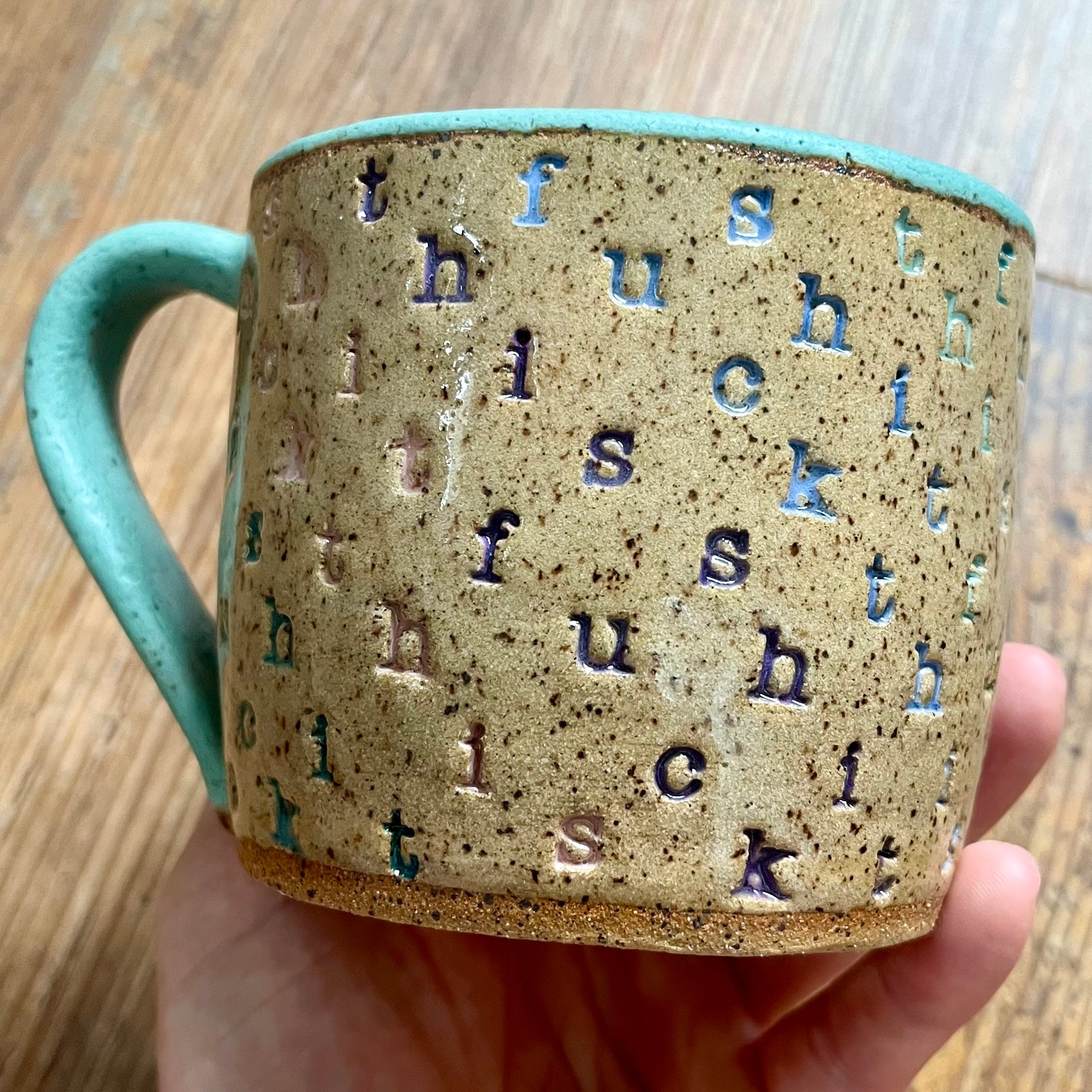 A mug stamped with "f*ck this sh*t" in diagonal stripes with a seaglass colored handle