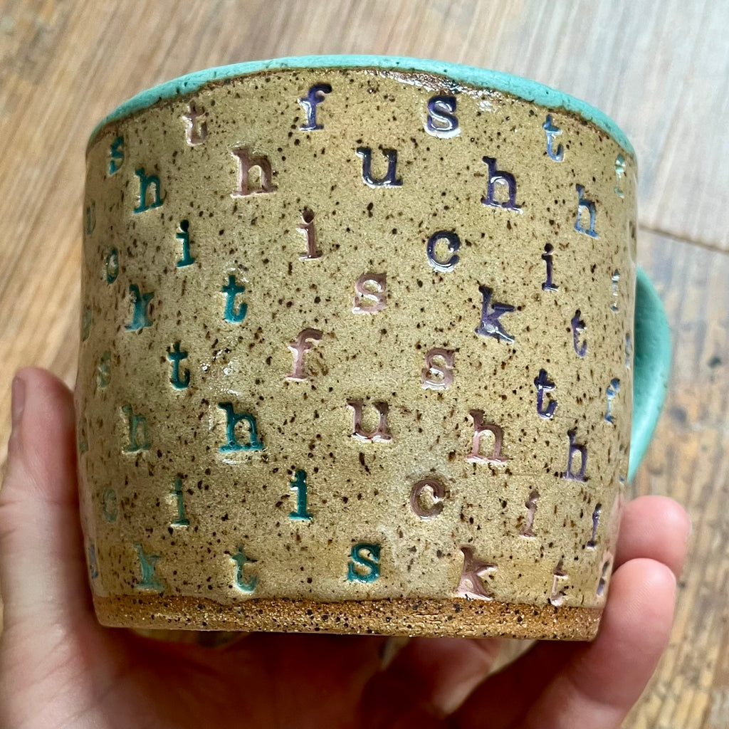 A mug stamped with "f*ck this sh*t" in diagonal stripes with a seaglass colored handle
