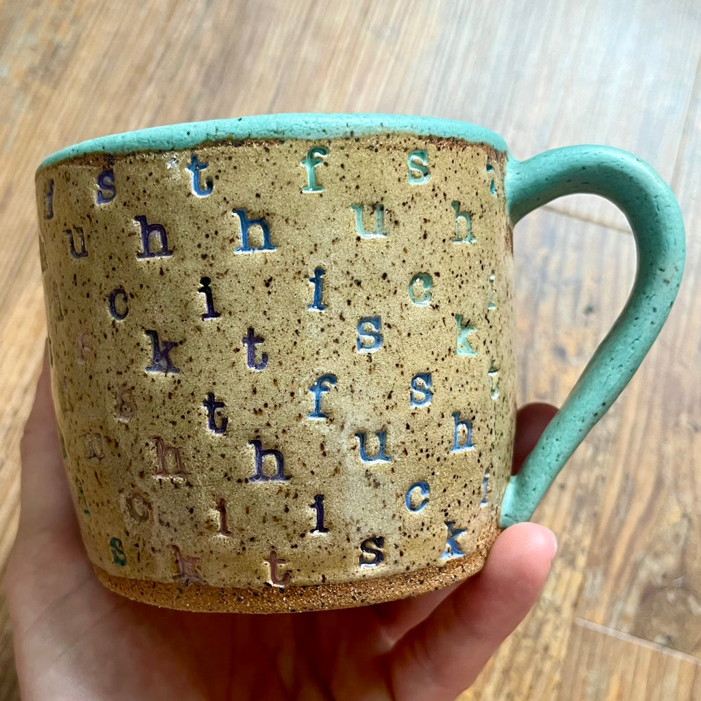 A mug stamped with "f*ck this sh*t" in diagonal stripes with a seaglass colored handle