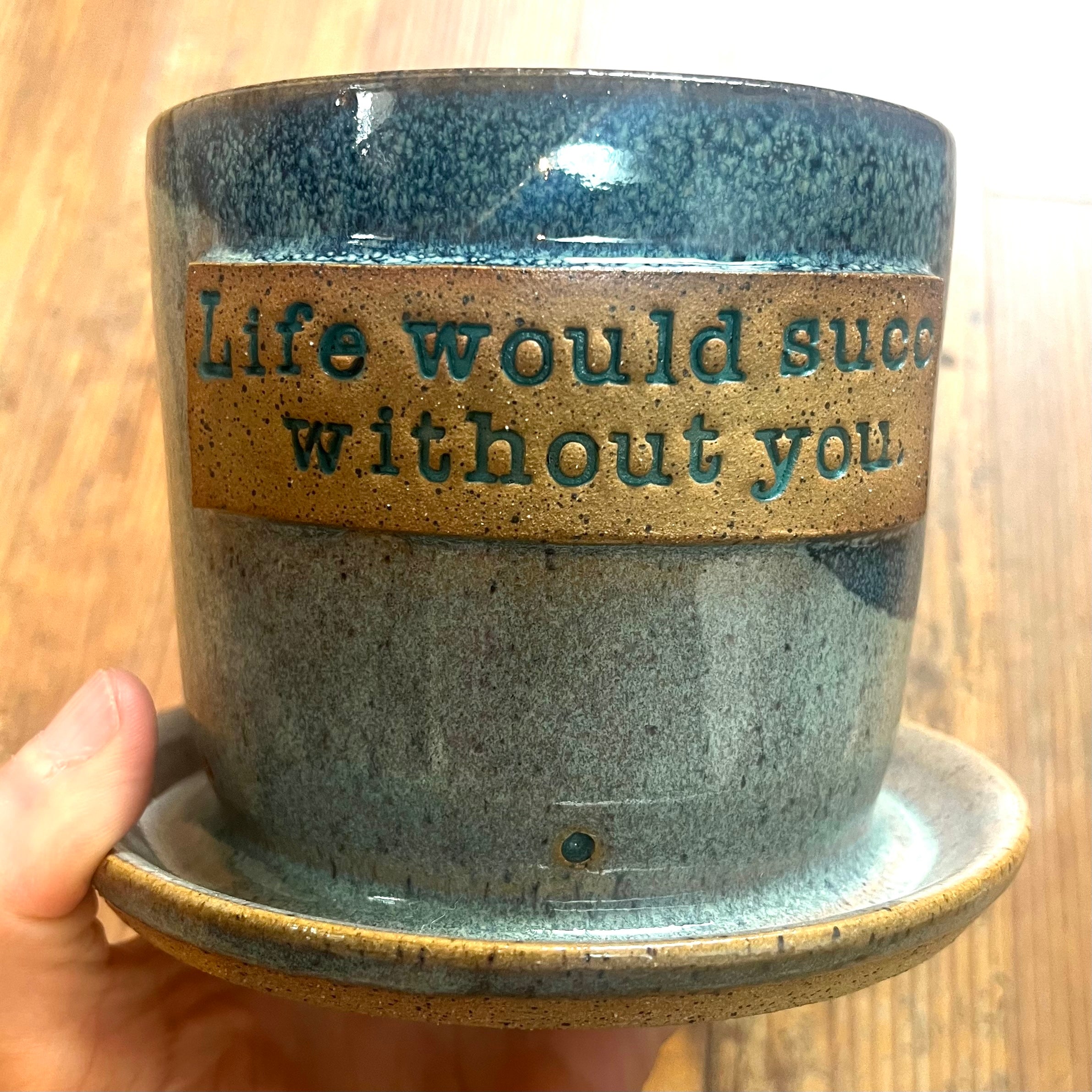 A handmade ceramic planter with an attached saucer, held by a hand. The planter features a speckled blue and gray glaze with a textured tan band around the middle containing the phrase 'Life would succeed without you' in a matching teal color.