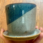 A handmade ceramic planter pot with an attached saucer, held in a hand against a wooden floor background. The pot features a diagonal split glaze design with a deep teal-blue top half and a speckled light gray-green bottom half, finished with a speckled tan rim.