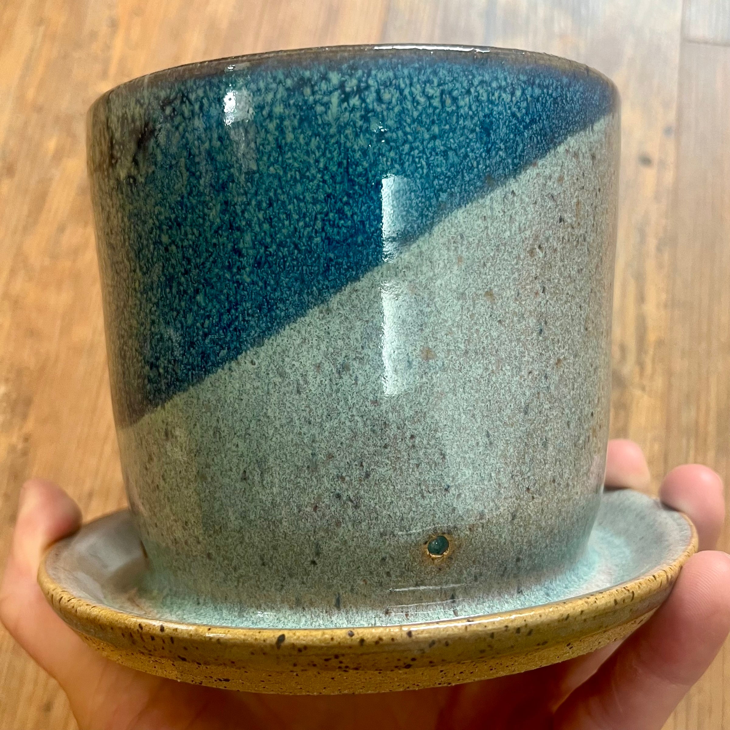 A handmade ceramic planter pot with an attached saucer, held in a hand against a wooden floor background. The pot features a diagonal split glaze design with a deep teal-blue top half and a speckled light gray-green bottom half, finished with a speckled tan rim.