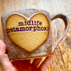 A light purple mug with blue specks featuring the phrase "midlife metamorphosis" in purple inside a clay heart