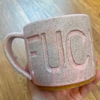 A hand holds a speckled light pink ceramic mug with the word 'FUCK' embossed on the side, set against a blurred wooden background.