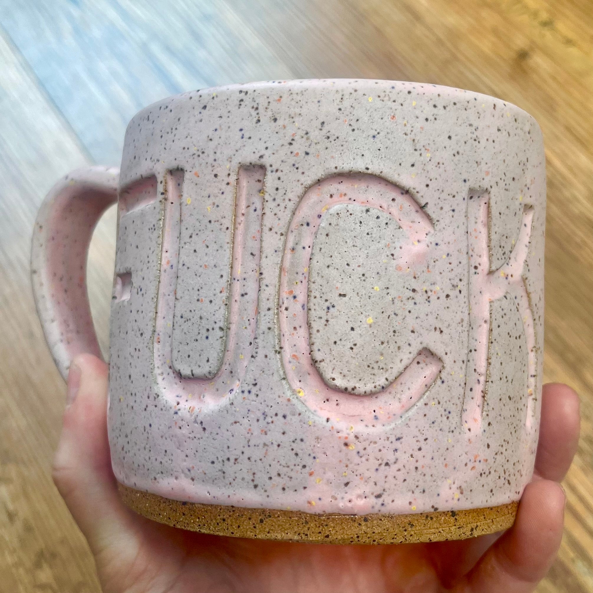 A hand holds a speckled ceramic mug with the word 'FUCK' embossed in large letters on the side. The mug has a light pink glaze with multicolored flecks, a cork-like tan base, and a handle on the left, shown against a blurred wood floor background.