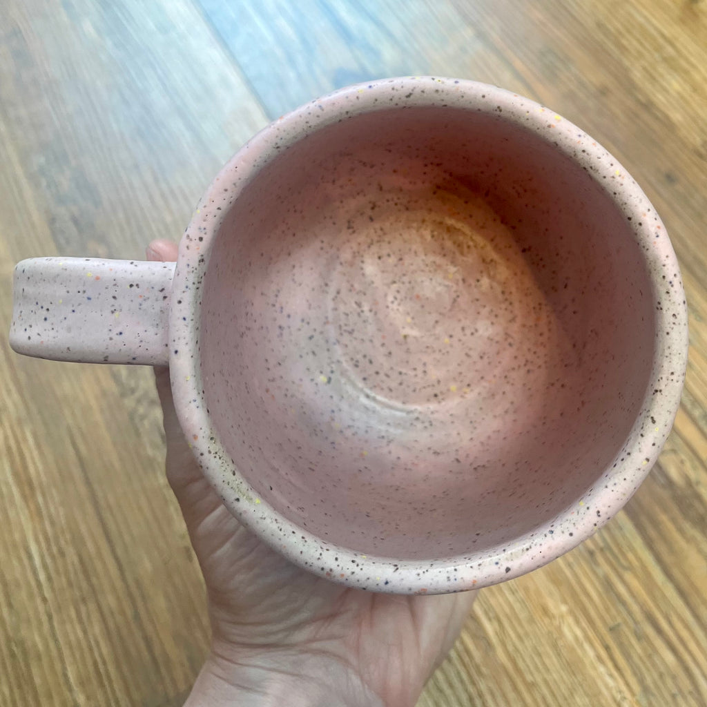 A hand holds a speckled pink ceramic mug with a handle, showing the interior of the cup against a wooden floor background.