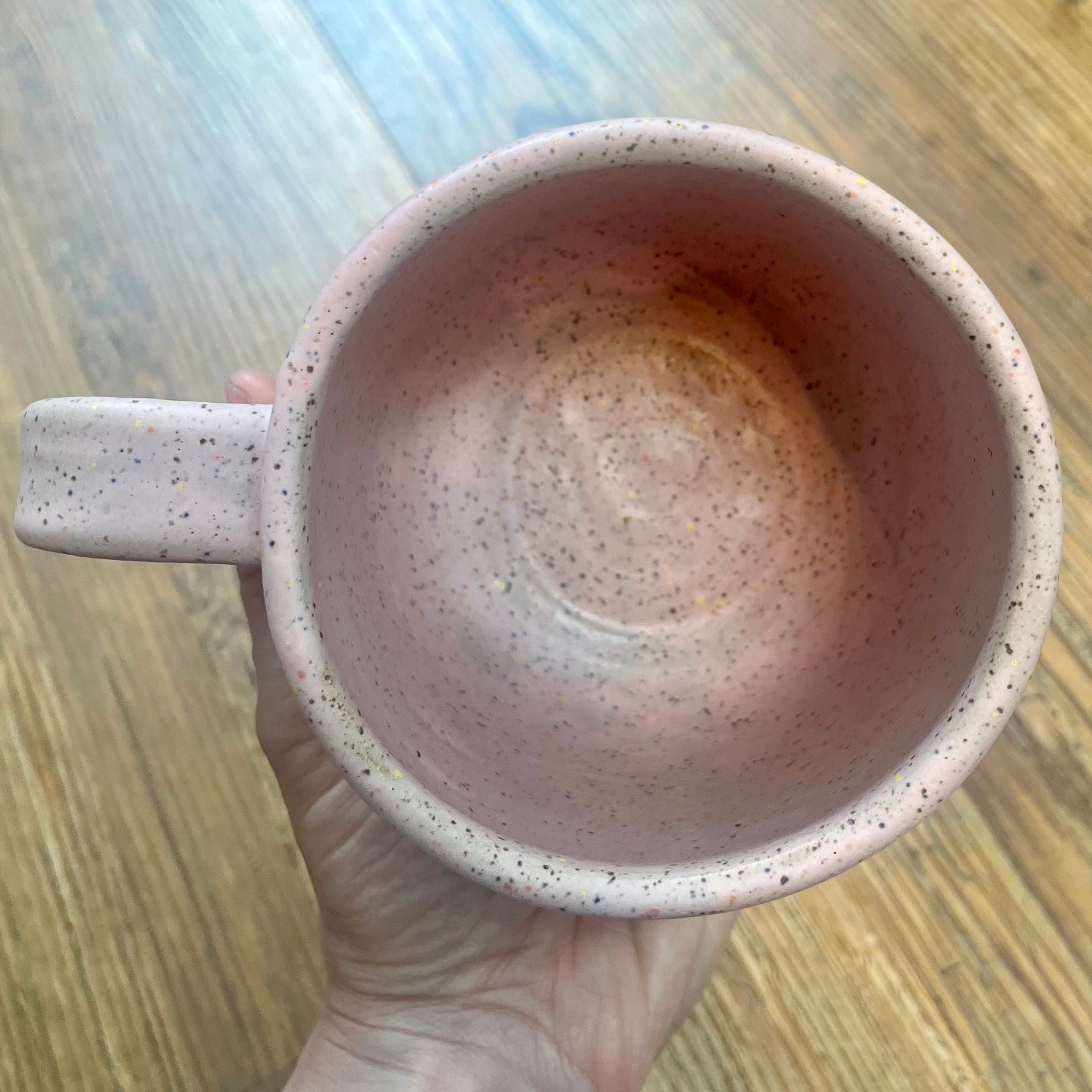 A hand holds a speckled pink ceramic mug with a handle, showing the interior of the cup against a wooden floor background.