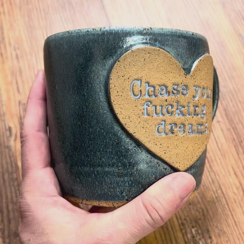 A video rotating around a greenish blue mug with the phrase "Chase your fucking dreams" inside a brown clay heart.