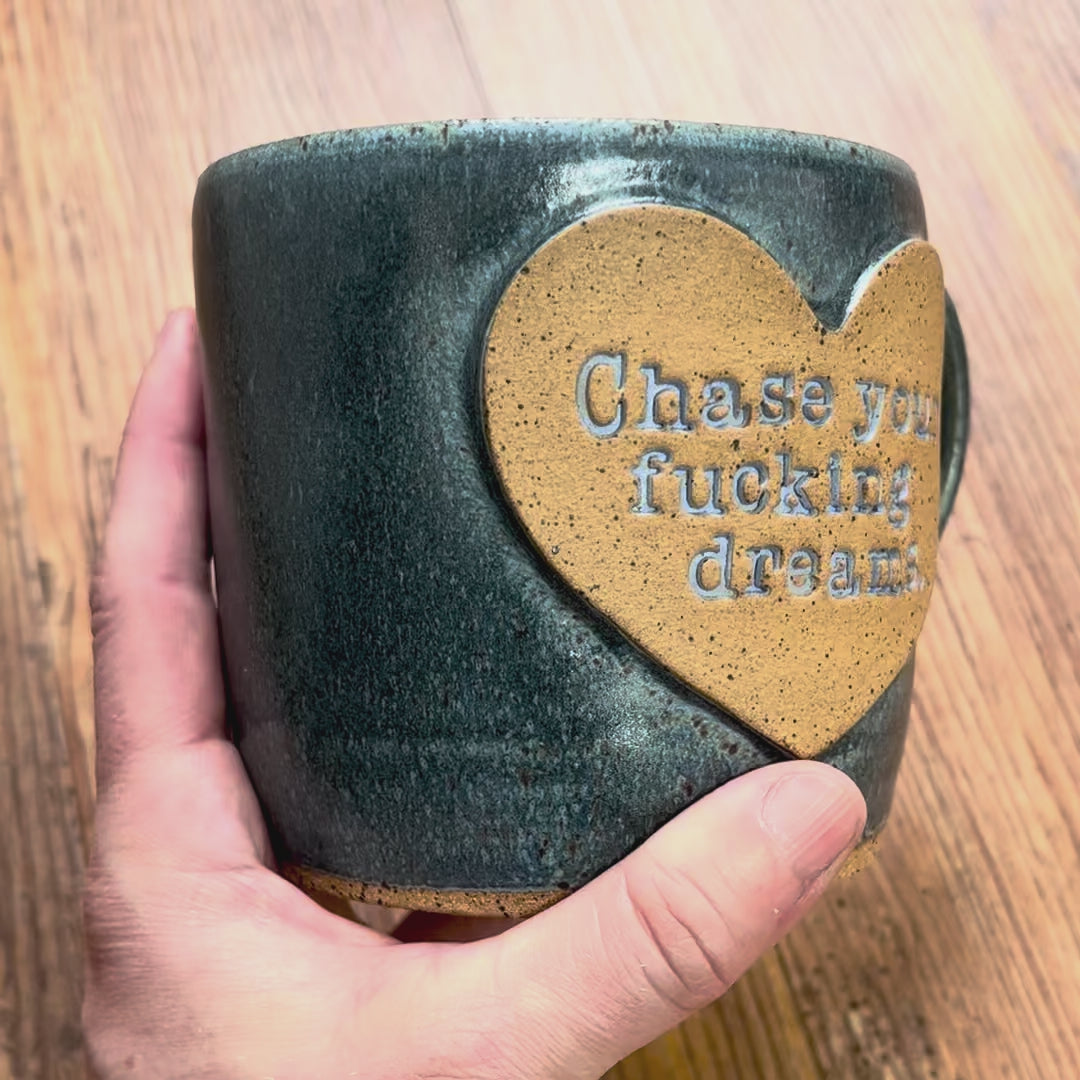 A video rotating around a greenish blue mug with the phrase "Chase your fucking dreams" inside a brown clay heart.