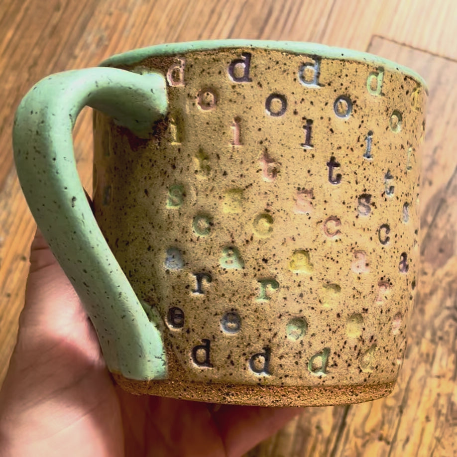 a video rotating around a mug with "do it scared" stamped in diagonal rainbow-colored stripes with a seaglass colored handle