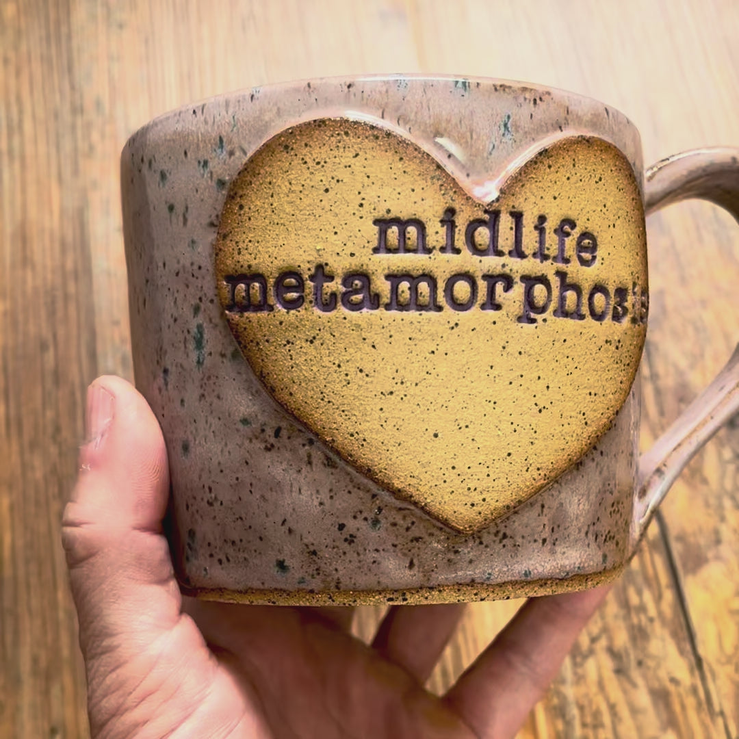A video rotating around a light purple mug with blue specks featuring the phrase "midlife metamorphosis" in purple inside a clay heart